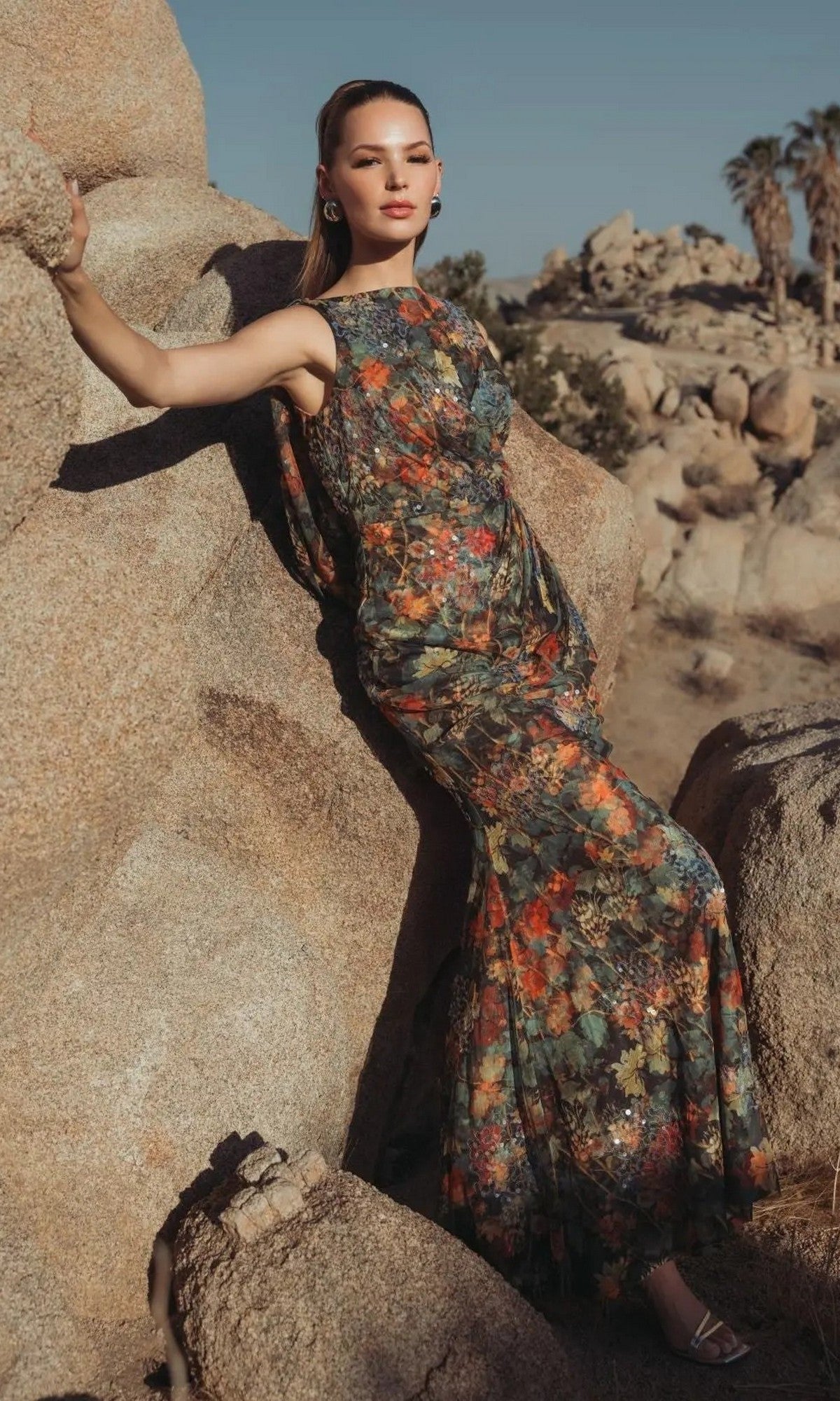 A woman in a sleeveless Mac Duggal 94263 Long Formal Dress in Charcoal Multi with floral patterns leans against a large rock in a rocky desert landscape, with palm trees visible in the background.